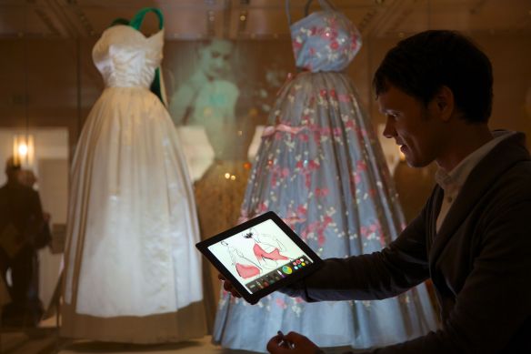 A visitor creates a fashion illustration using an interactive app with dresses worn by HM The Queen in the background.